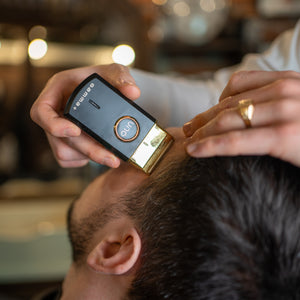 A close-up of a barber using gamma+ uno shaver black and gold electric hair clipper on a client's head in a barbershop setting.