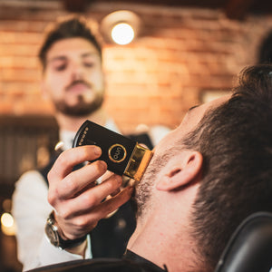 A close-up lifestyle shot of a barber using a black and gold electric Uno Professional Single Foil Shaver on his beard, with a barber visible in the blurred background in a warm, brick-walled barbershop setting.