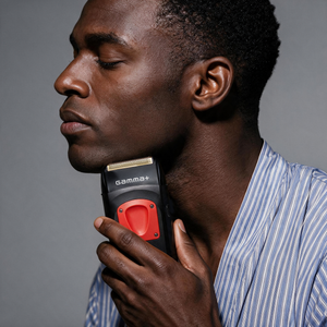 A close-up of a man holding a Gamma+ electric shaver against his neck, shown in profile. The man is wearing a blue and white striped shirt, and the background is a solid gray studio backdrop.