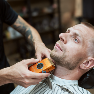 A man in a barber chair while a barber uses an orange and blue Braun beard trimmer on his beard. The setting is a barbershop with a blurred background.