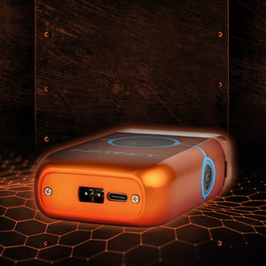 An orange metallic Instinct vape device with blue accents, shown from a rear three-quarter angle against a dark wood panel background with a glowing orange hexagonal grid pattern.