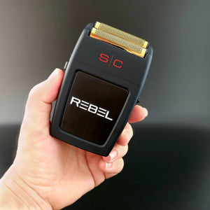 A hand holding a Rebel Professional Double Foil Shaver with a gold-colored shaving head and red 'S/C' markings, featuring the brand name 'REBEL' in white on the front.