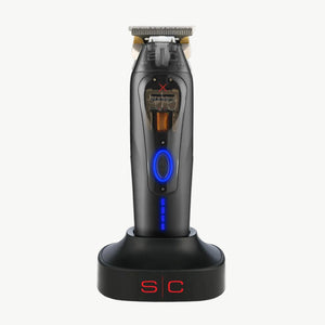 360 X Jeezy trimmer standing upright on a matching black charging dock against a plain white background. The shaver has a blue circular control button, a small display screen, and metallic silver blades at the top.