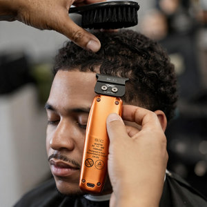 A close-up of a person getting a haircut with a barber using an orange and black SIG electric clipper on curly dark hair. A hand is brushing the hair while the other holds the clipper, with a blurred barbershop background.