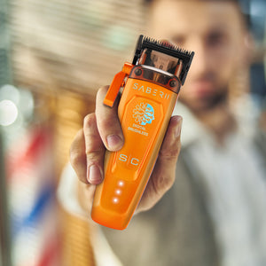 A close-up of a person holding an orange Saber 2 hair clipper against a blurred indoor background.
