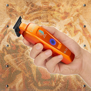 A hand holding an orange pet grooming clipper against a textured, rusted metal background with small metal studs.