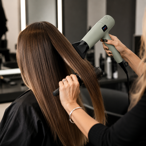 A salon setting with a stylist using a light sage green hair dryer on a client's long, straight brown hair. The client is seated with a black salon cape on, and the background shows blurred salon fixtures.