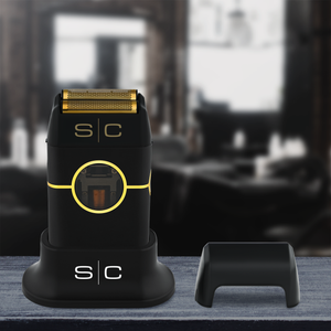 Instinct Metal Professional Double Foil Shaver with gold accents and a matching charging dock, shown alongside a small black accessory on a dark wooden surface with a blurred indoor background.