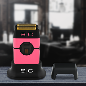 Instinct Metal Professional Double Foil Shaver with a gold-colored shaving head, shown standing on a black charging base with its cap off beside it. The background is a blurred indoor setting.